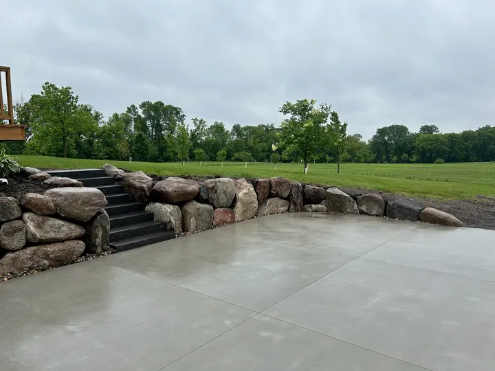 Boulder retaining wall with stone steps beside a new concrete patio, open lawn beyond