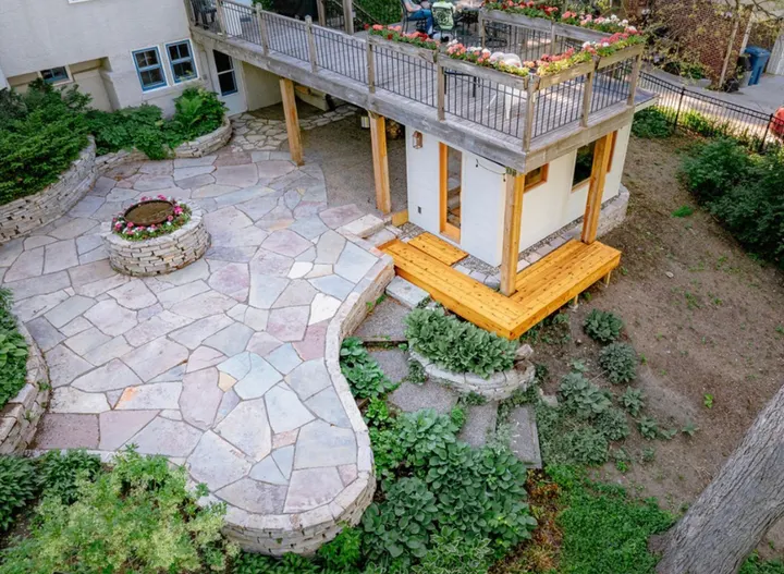 Drone view of a Chilton flagstone patio with a round stone fire pit, set below a raised deck and bordered by boulder walls and plantings