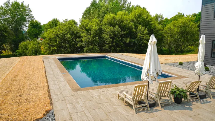 Concrete pool deck with a paver border, lounge chairs, and umbrellas at the far end, open field beyond