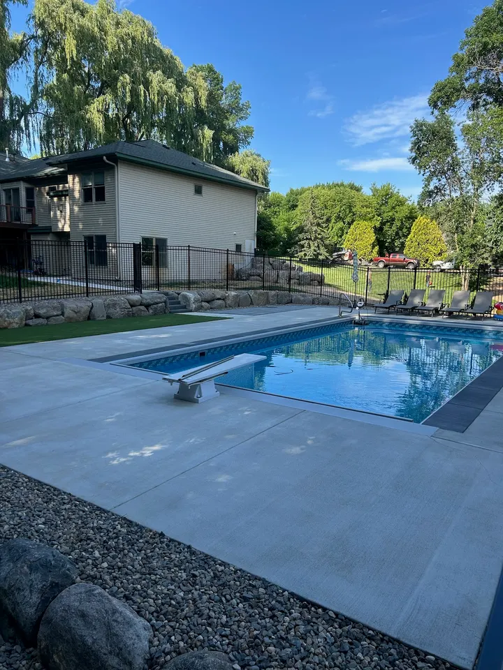 Light gray concrete pool deck with a diving board and black fence, house and trees in the background