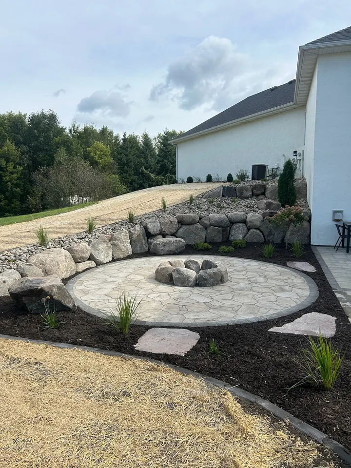 Round paver patio with a stacked boulder fire ring, a boulder retaining wall on the uphill side, stepping stones through mulch beds, and a white house behind